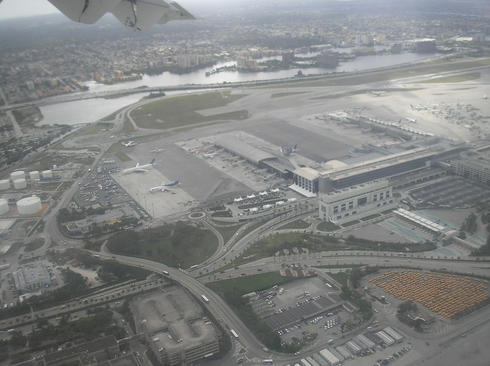 Aerial photograph of Miami International Airport on approach: the Central Terminal and concourses, apron with parked aircraft, the landside roadway loops, parking structures, and the Miami skyline in the distance