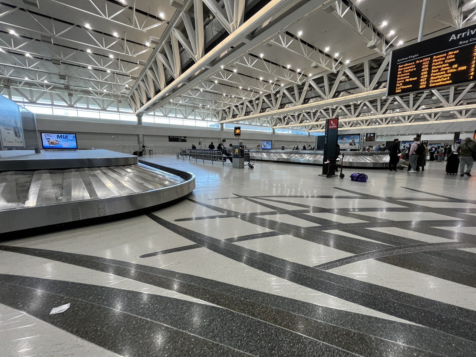 Interior photograph of the international arrivals baggage claim hall at Miami International Airport: a carousel in the foreground, exposed white roof trusses, an arrivals board listing Latin American and European flights, and passengers waiting at the far carousels
