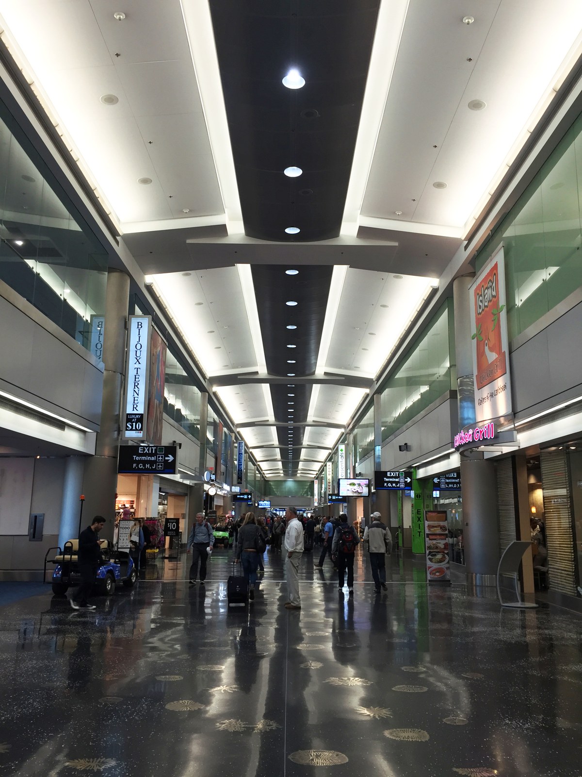 Interior photograph looking down the long Concourse D retail corridor at Miami International Airport: a vaulted lit ceiling, storefronts on both sides, exit signage to Terminals F, G, H, and J, and passengers walking along the polished floor