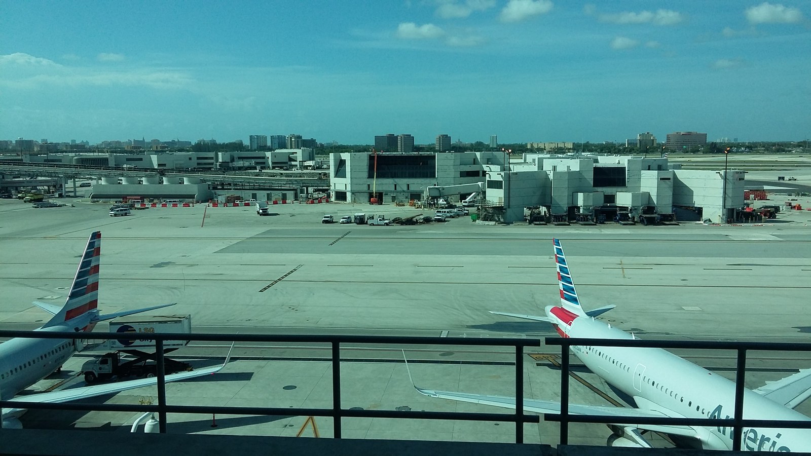 Photograph from the Skytrain platform looking across the apron toward Concourse E: two American Airlines regional jets at gates, catering trucks, the concourse pier and jet bridges, and the Miami skyline under a blue sky