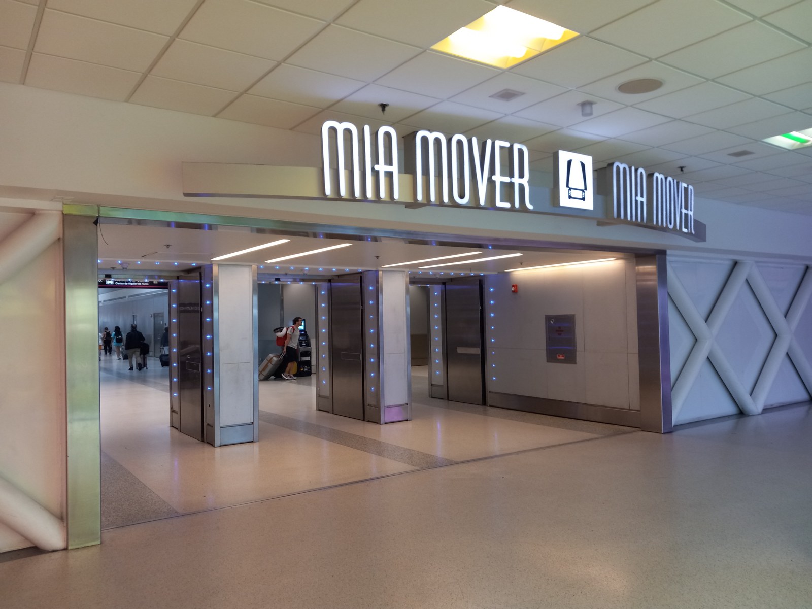 Photograph of the MIA Mover station entrance inside the terminal: an illuminated MIA MOVER sign above fare-style gates with blue LED edges, terrazzo floor, and passengers with luggage passing through