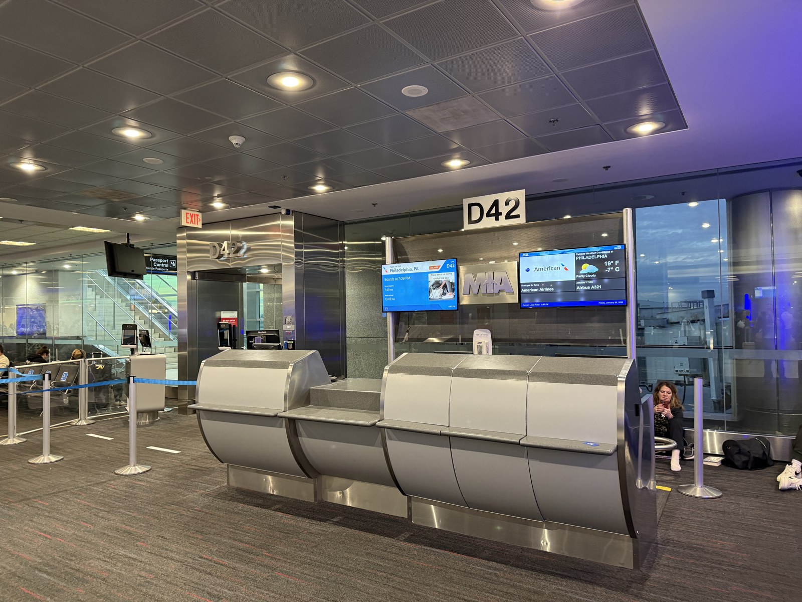 Interior photograph of gate D42 in Concourse D at Miami International Airport in January 2026: the curved gate desk, MIA lettering on the back wall, two flight-information screens for an American Airlines departure, stanchions, and floor-to-ceiling glass onto the apron
