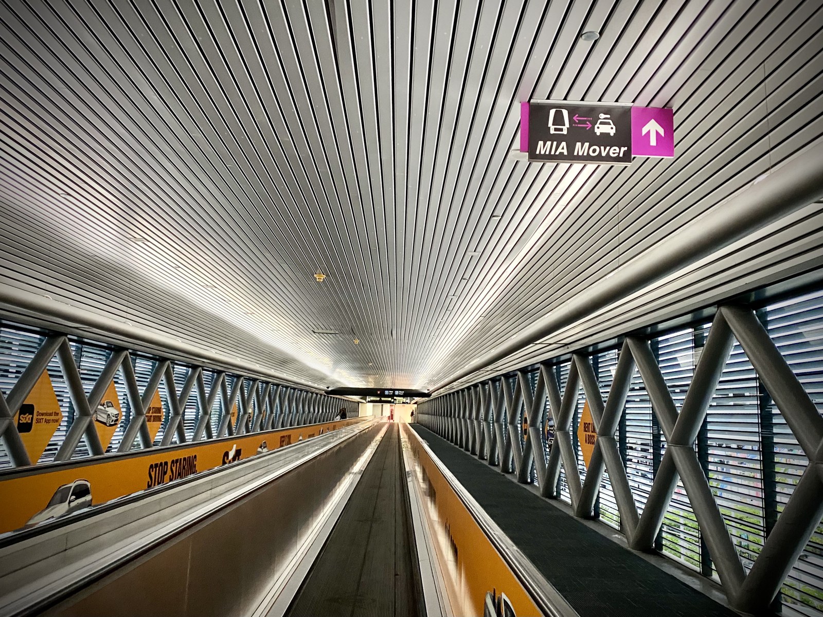 Photograph inside the MIA Mover connector walkway: moving sidewalks under a ribbed metal ceiling, a magenta MIA Mover sign with a train icon, and diagonal steel bracing along glass walls
