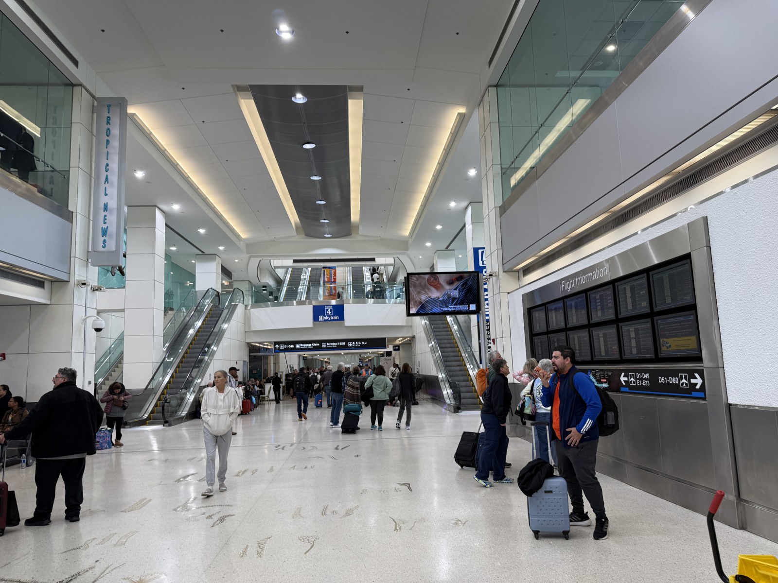 Interior photograph of the Concourse D hall at Miami International Airport in January 2026: terrazzo floor, escalators up to Skytrain Station 4, a wall of flight-information screens, directional signage to gates D1 to D49 and D51 to D60, and passengers with luggage