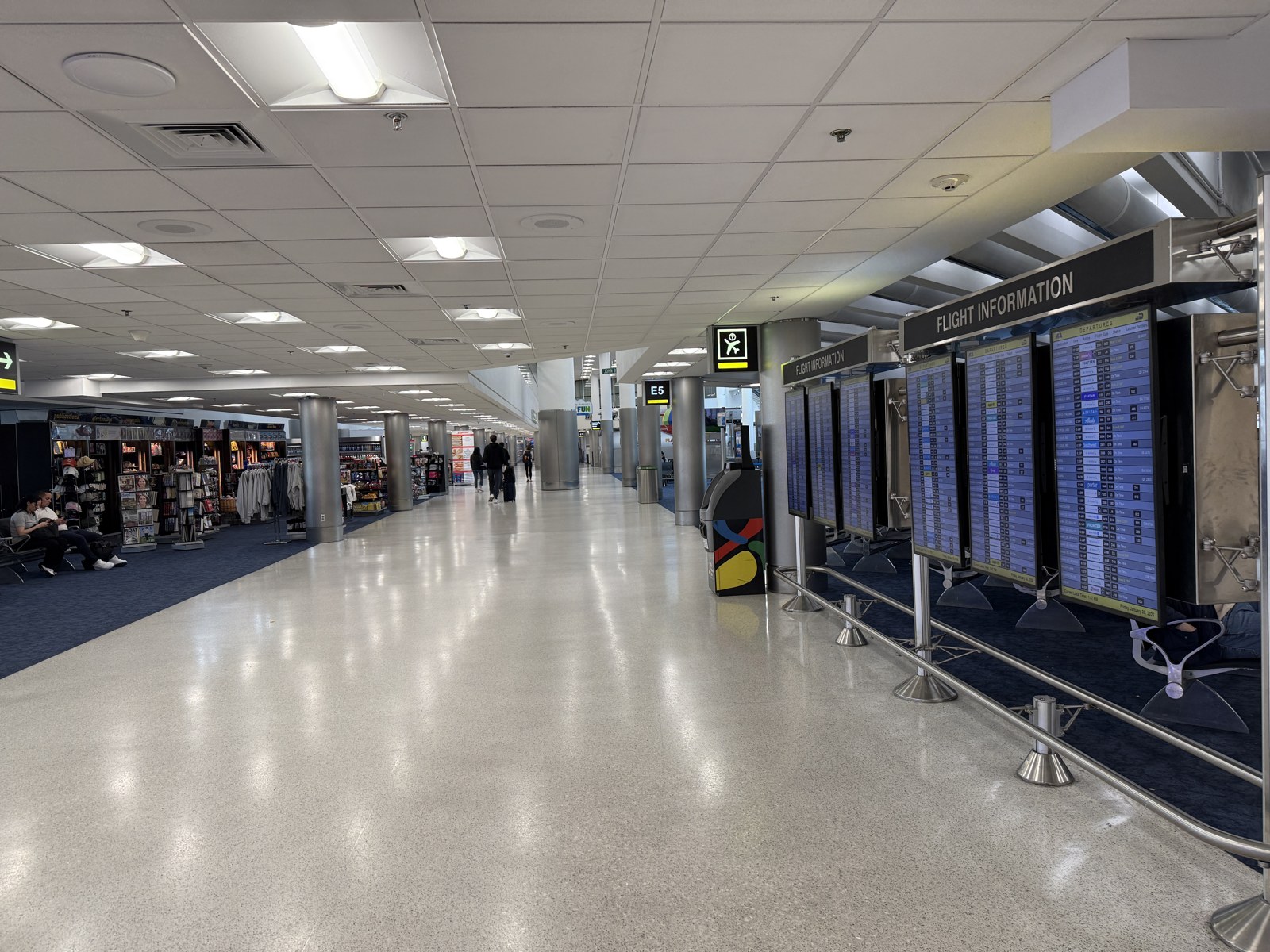 Interior photograph of Concourse E at Miami International Airport in January 2026: a bank of flight-information screens on the right, a newsstand and shops on the left, and a wide terrazzo corridor toward gate E5